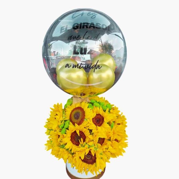 Arreglo floral de girasoles amarillos brillantes en base blanca con un globo transparente con la frase "Eres el girasol que le da luz a mi vida" y globos dorados dentro.